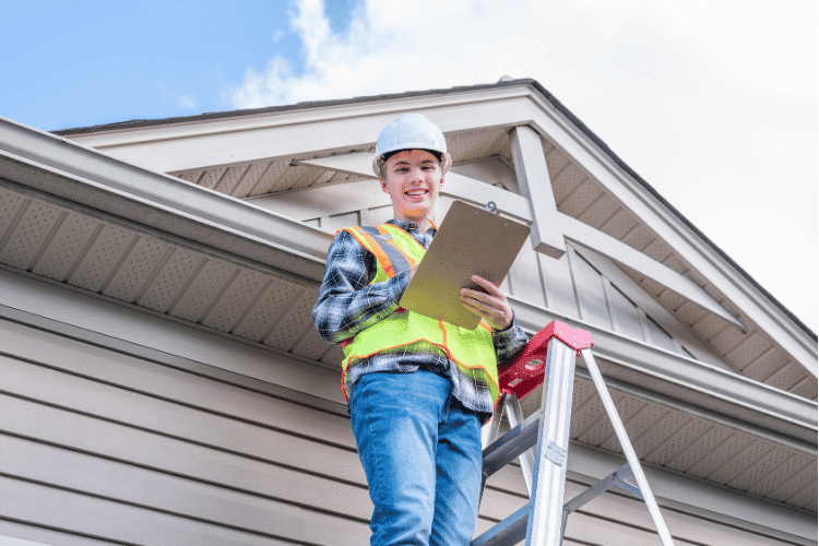 A smiling Home Inspector standing on a ladder and providing inspection to the roof of a house